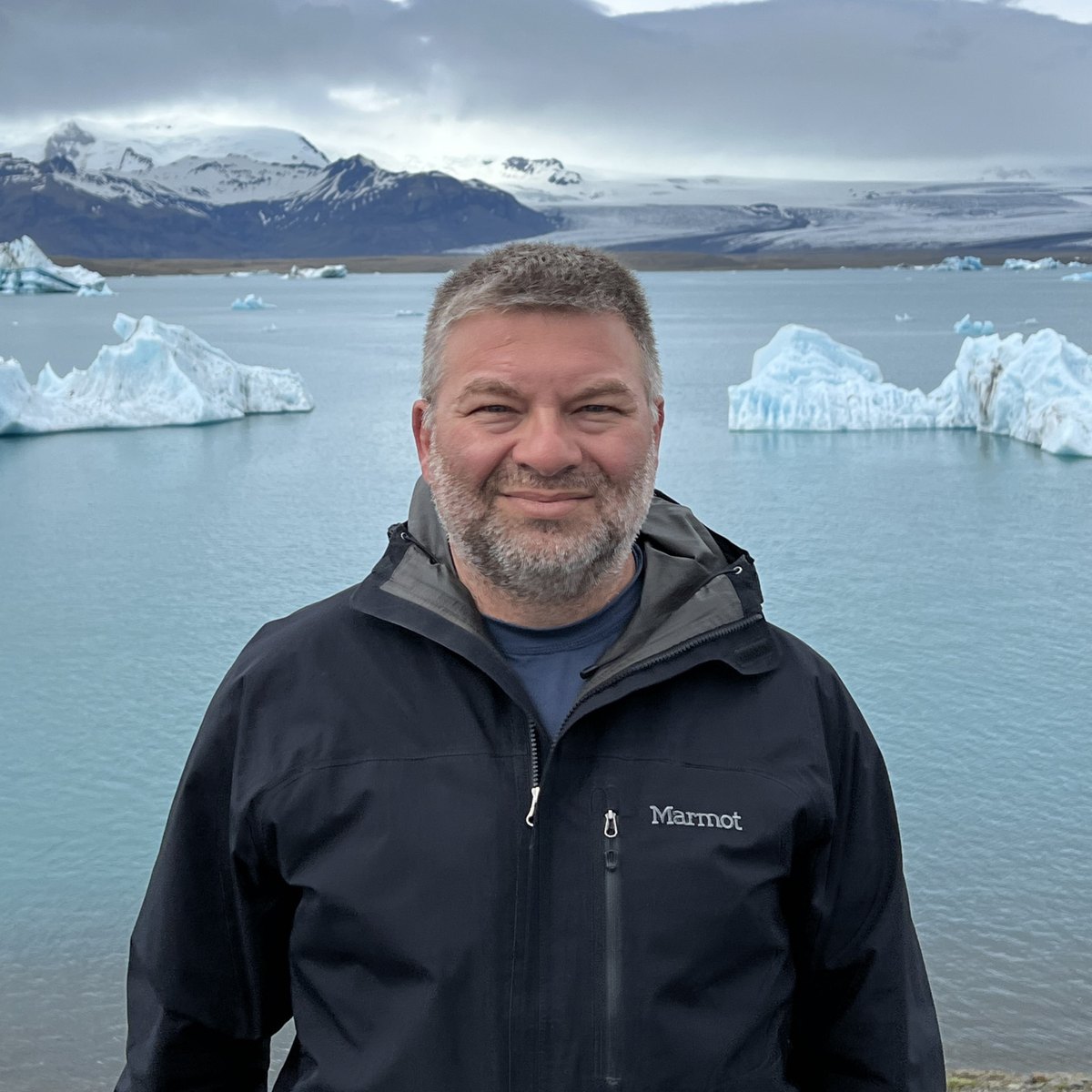 Chris Commisso standing beside a glacial lagoon in Iceland, with icebergs and a snow-covered mountain behind him.