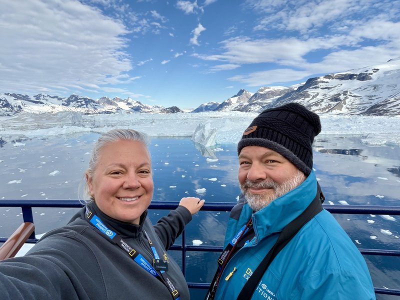 Erika and Chris Commisso on an expedition ship in Greenland, with icebergs and snow-covered mountains behind them.