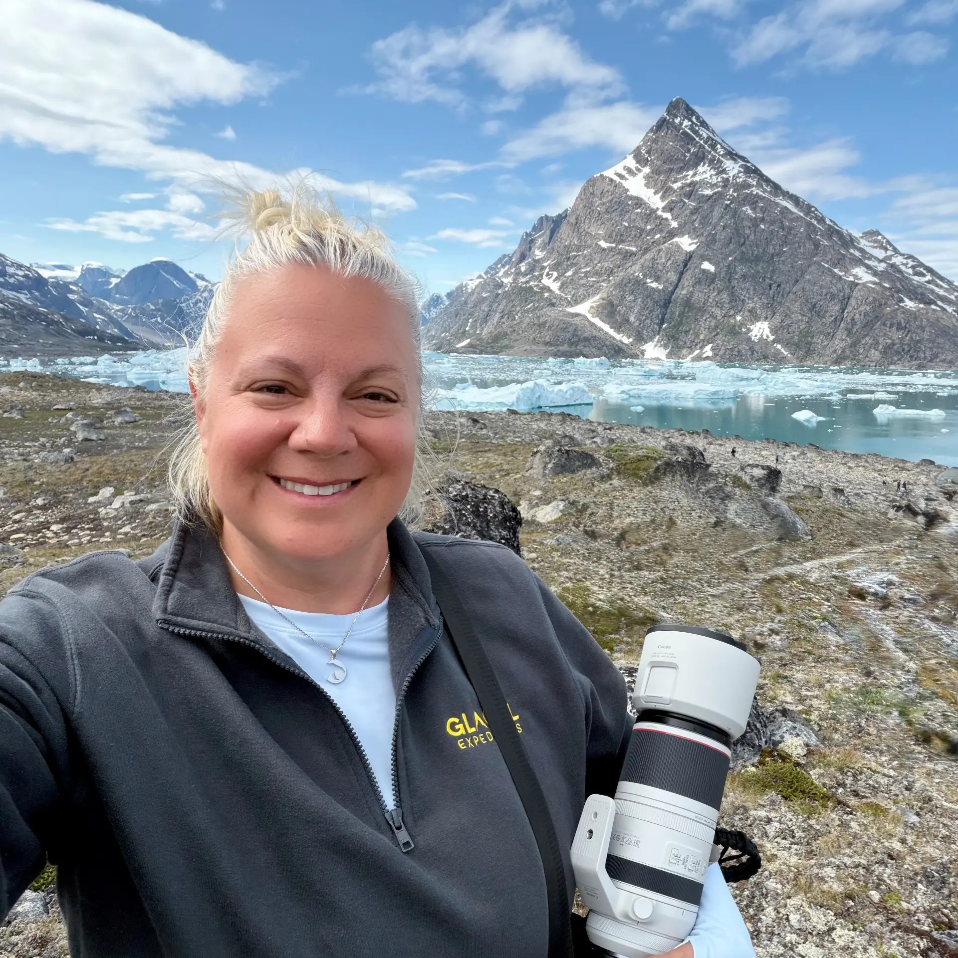 Erika Commisso smiling in expedition clothing with a camera beside a glacial lake with floating ice and a snow-covered mountain in Greenland.