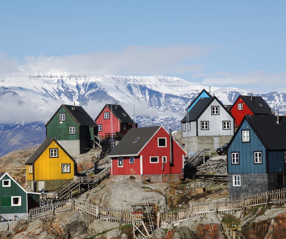 Brightly painted red, yellow, green and blue wooden houses on bare rock below a snow-covered mountain range in Greenland.