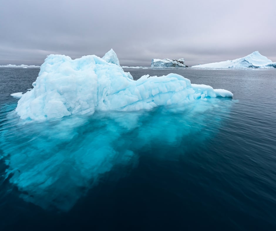 A luminous turquoise iceberg floating in calm grey Greenlandic water under an overcast sky.