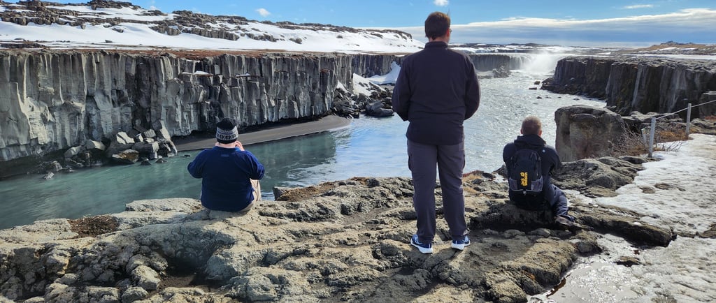 Travelers on basalt cliffs overlooking a wide waterfall pouring into a volcanic canyon in Iceland, snow still on the ground.