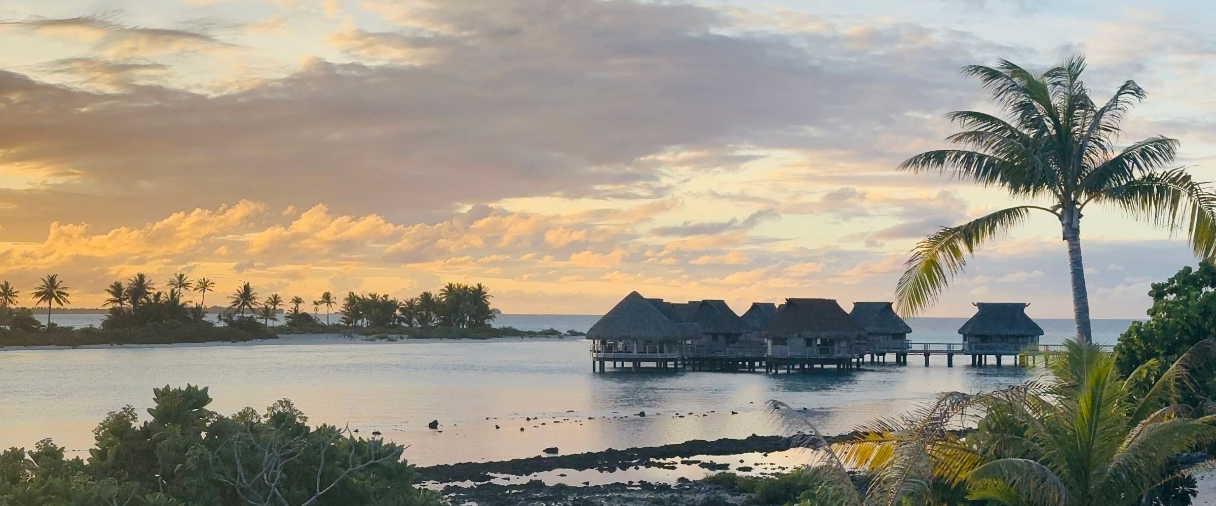 Sunset over a Tikehau lagoon with thatched overwater bungalows and coconut palms silhouetted against an orange sky.