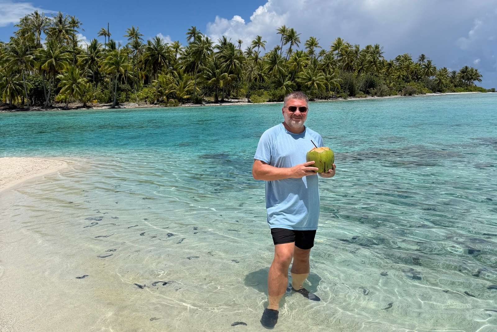 Chris Commisso standing in clear shallow water at Motu Mahana, holding a fresh coconut, with a palm-lined shore behind.