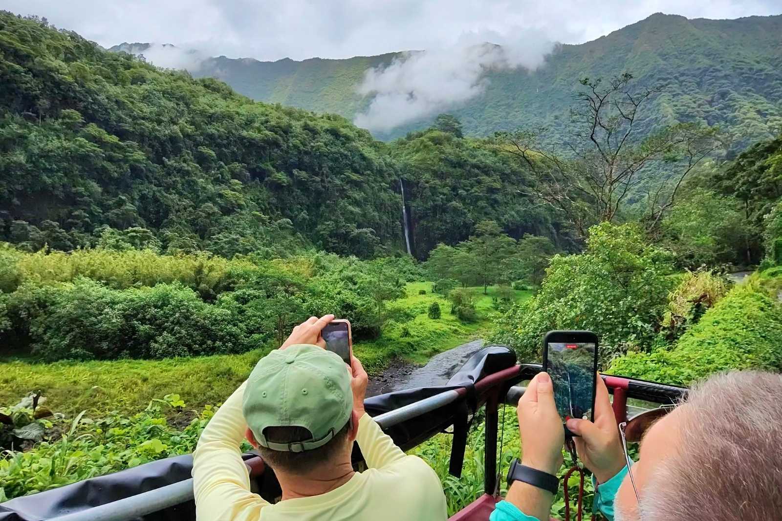 Passengers in an open-air 4x4 photographing a waterfall in the green interior of Tahiti Nui, with cloud on the ridges.