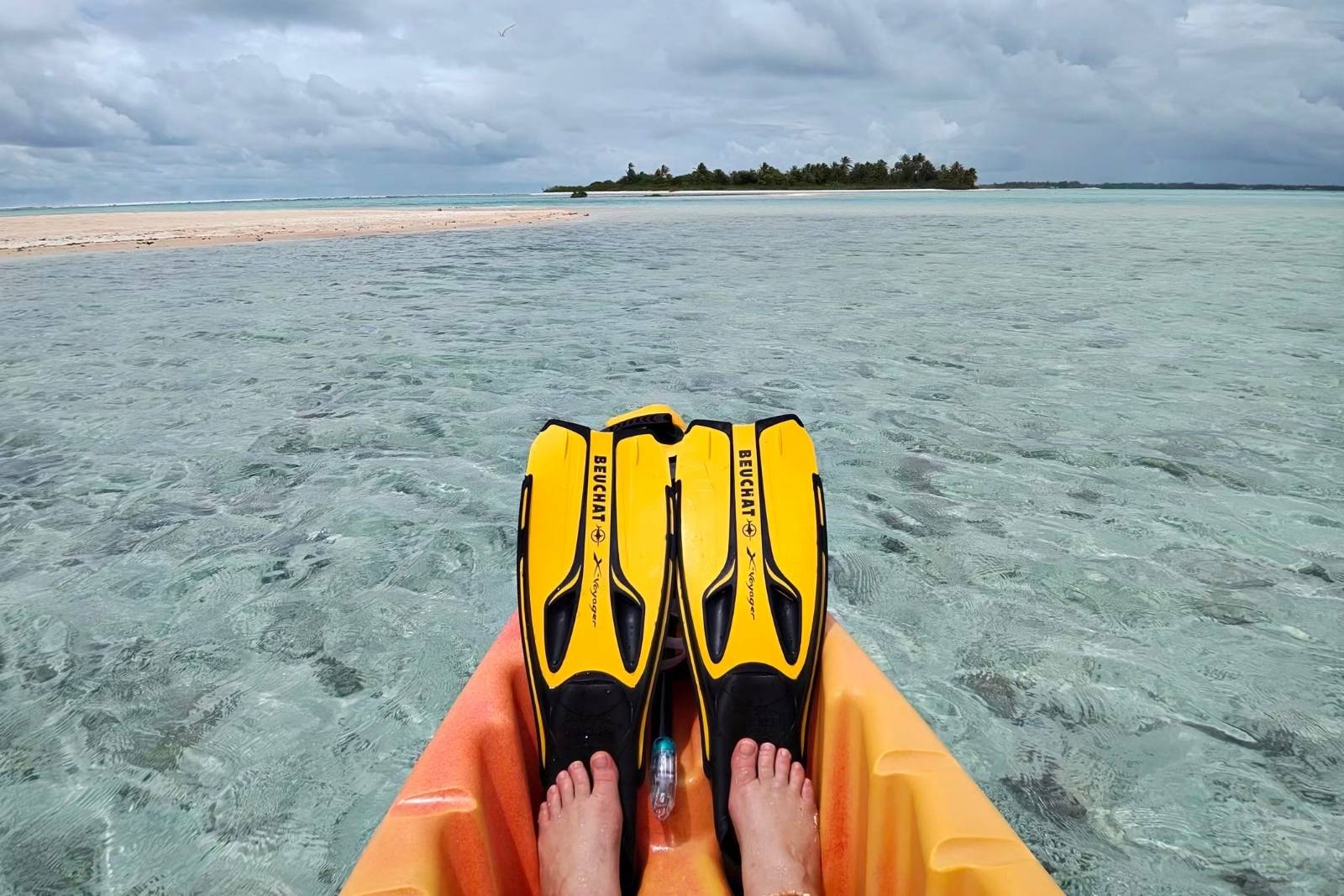 The view forward from a kayak across clear shallow water at Tikehau, with snorkel fins on the bow and a palm-covered motu ahead.