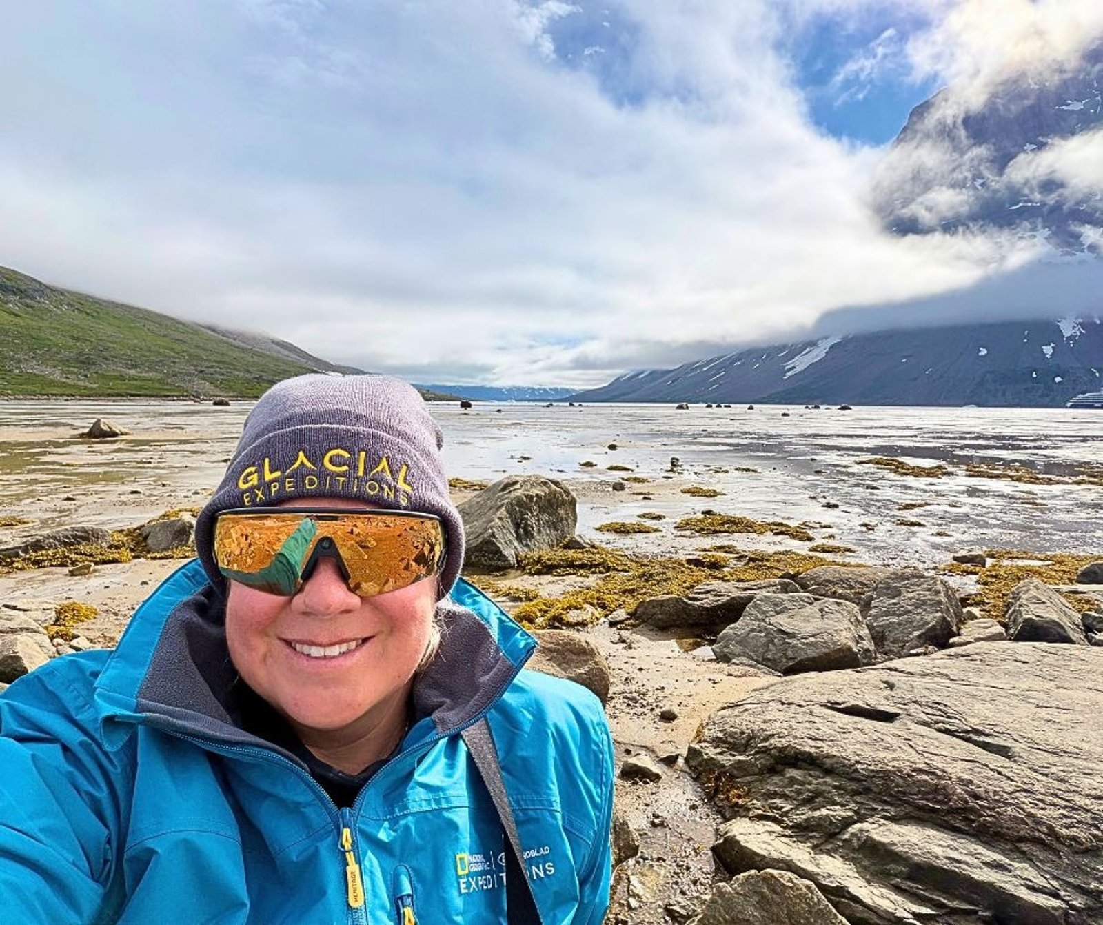 A person in a Glacial Expeditions beanie and sunglasses on a rocky Arctic shoreline with mountains behind.