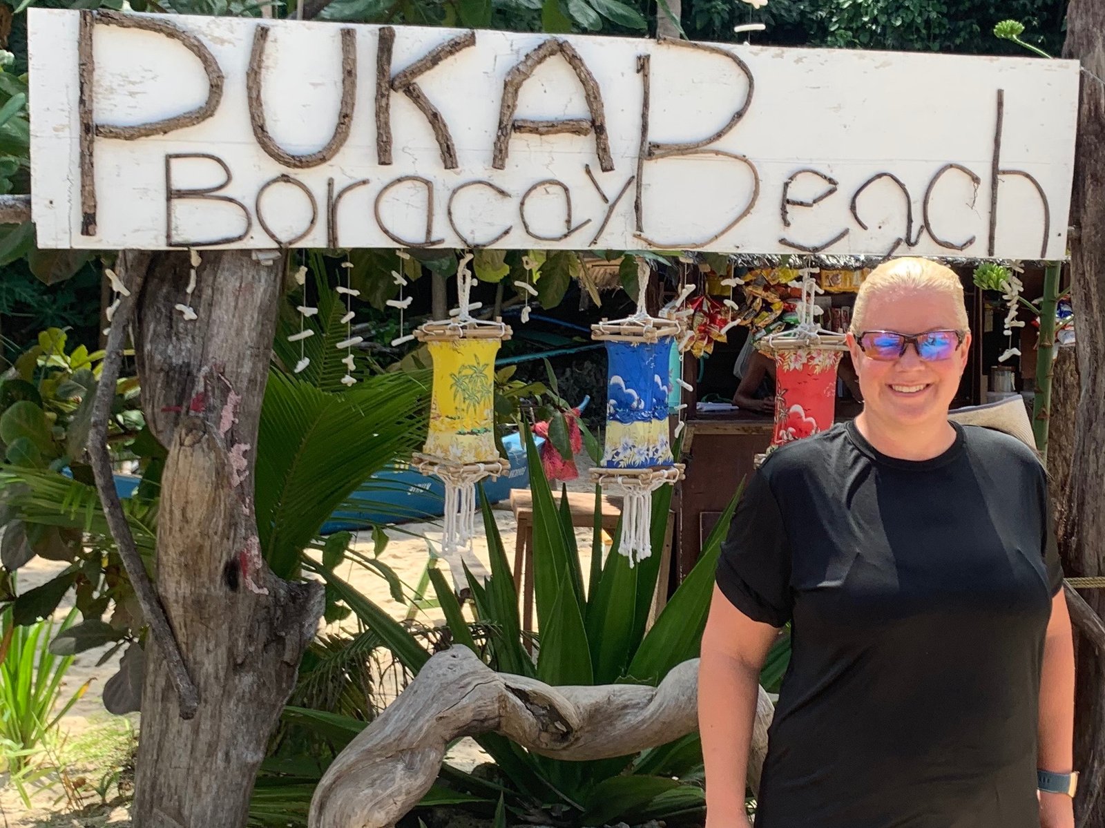 Erika Commisso beside a hand-lettered Puka Boracay Beach sign hung with painted paper lanterns and shell strings.