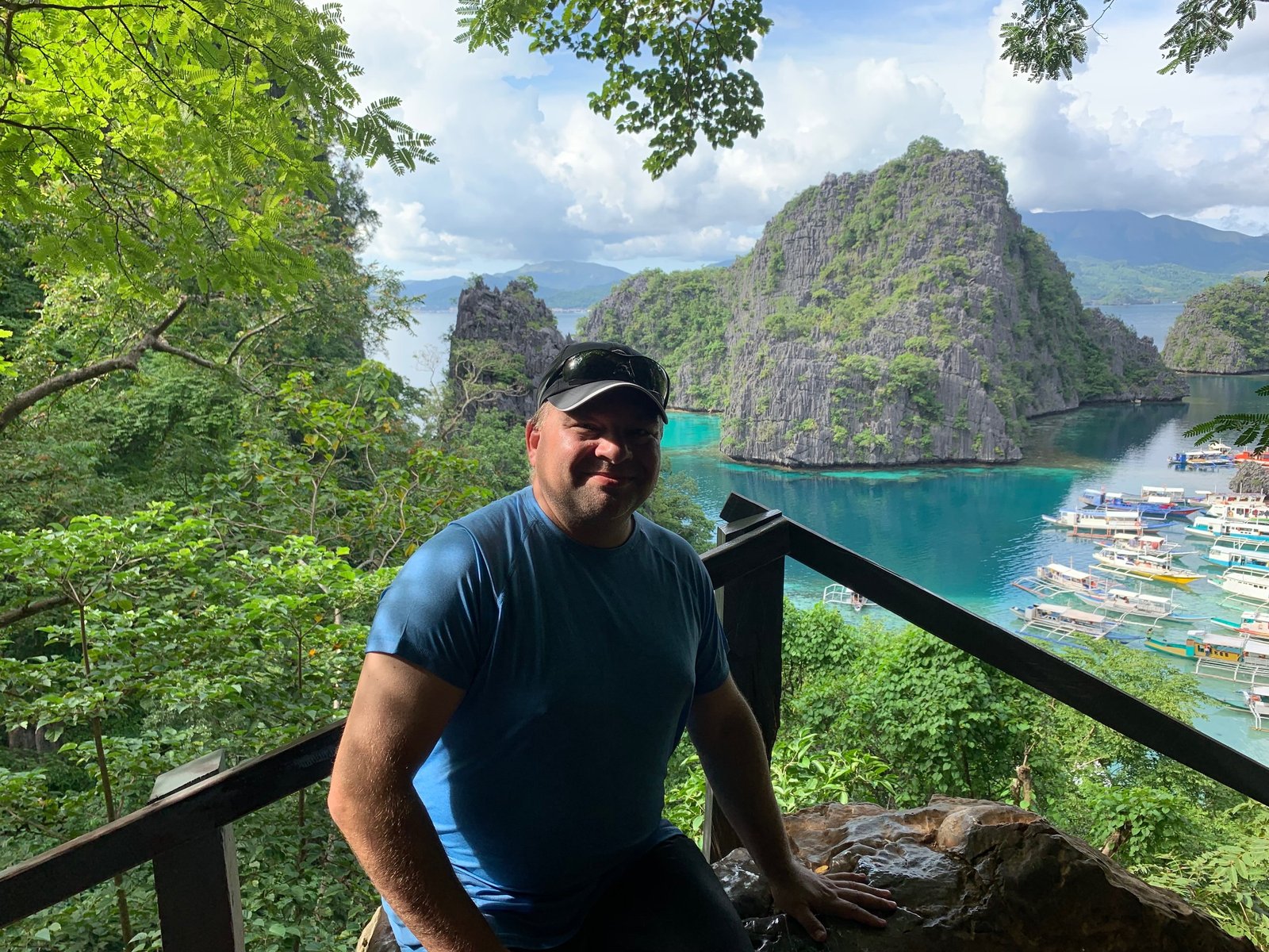 Chris Commisso at a jungle viewpoint above a turquoise Philippine bay ringed by limestone karst islands and moored outrigger boats.