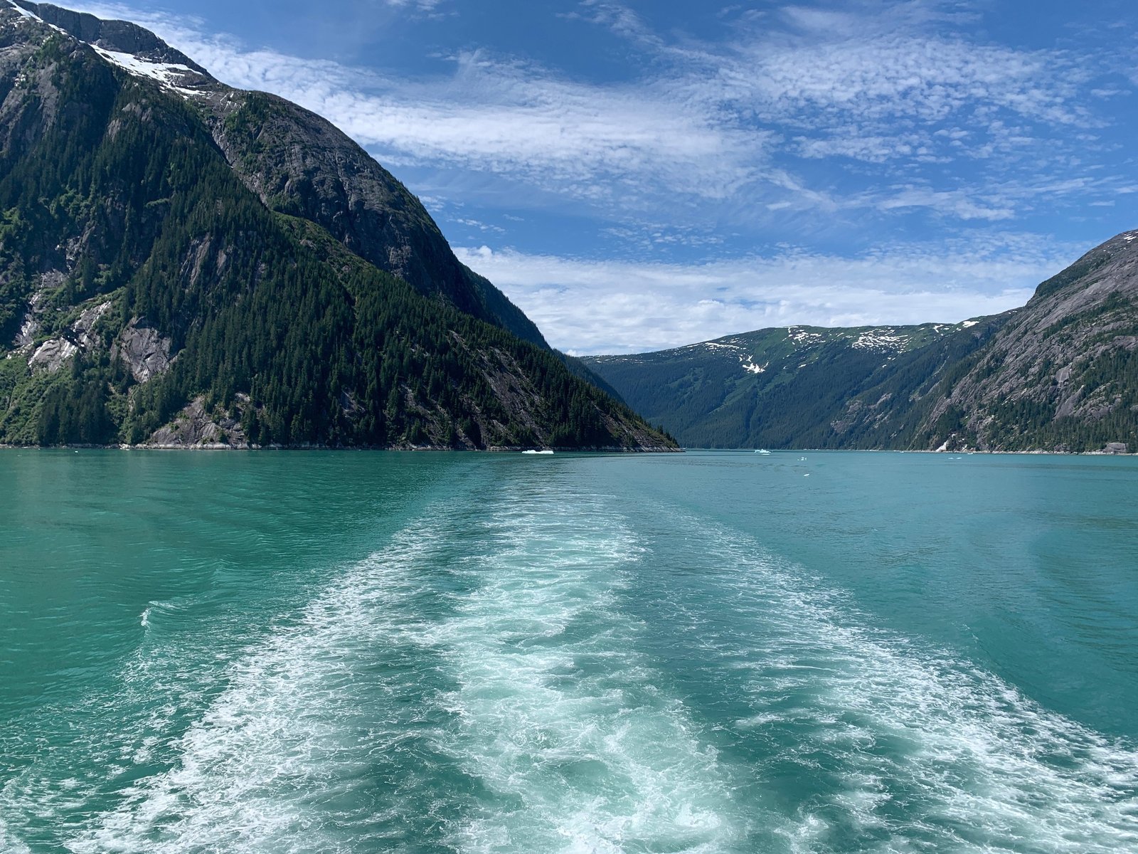 The wake of an expedition ship trailing through a turquoise Alaskan fjord between forested granite walls.