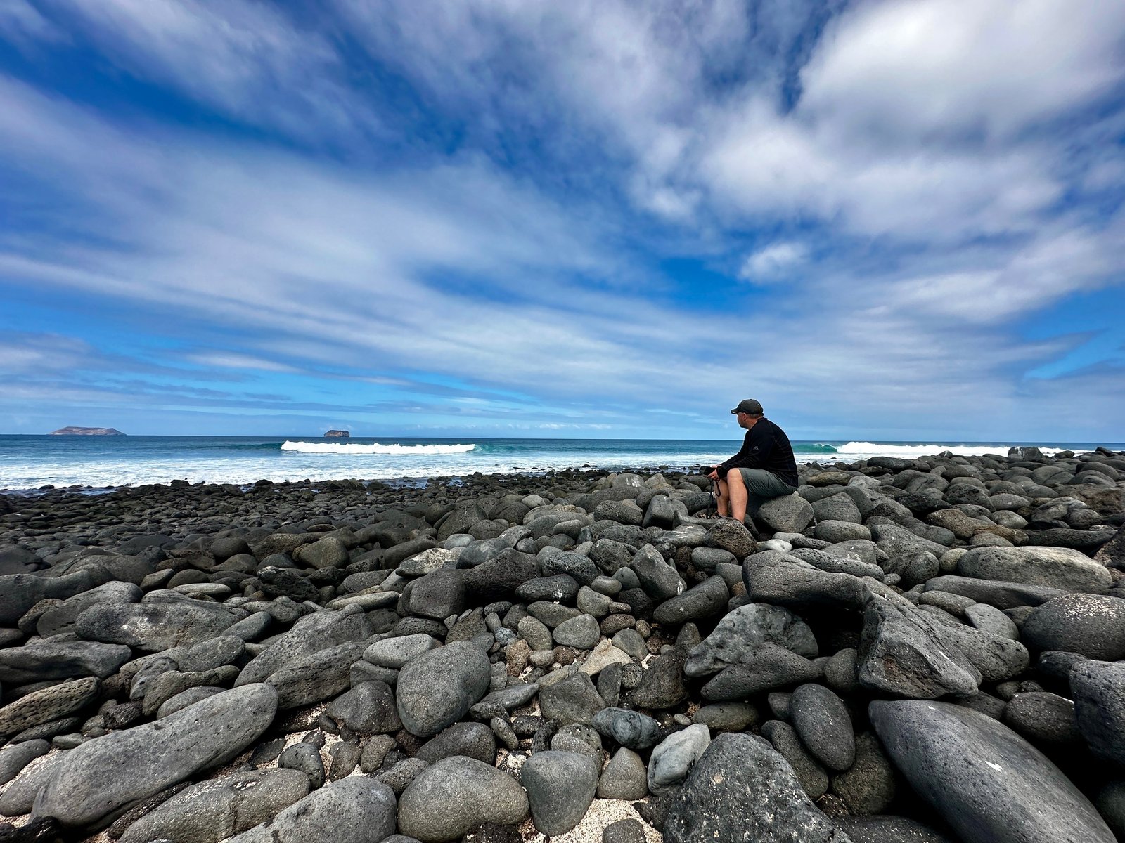 A traveller sitting on a boulder-strewn volcanic shoreline in the Galapagos, surf breaking beyond and small islets on the horizon.