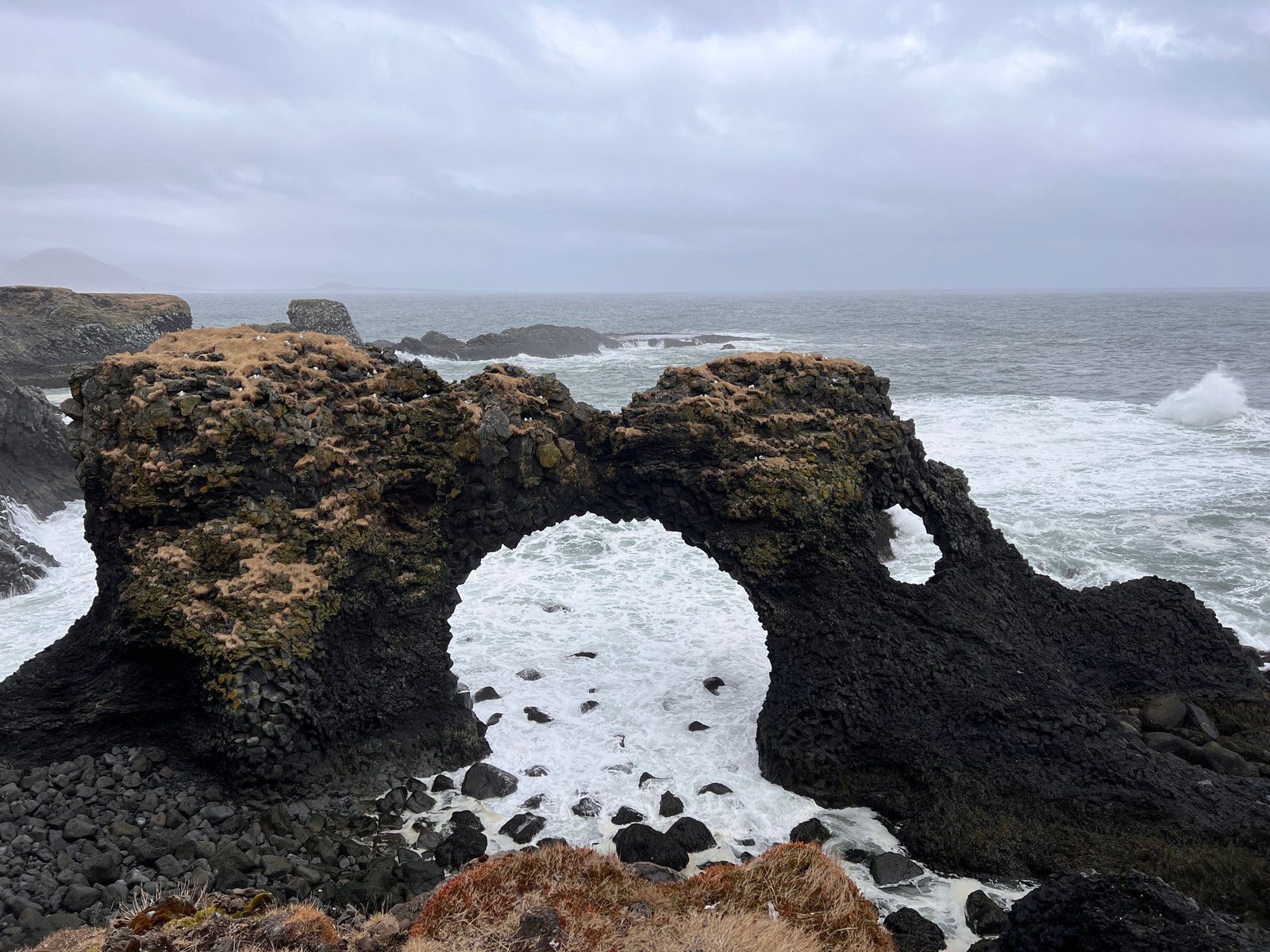 A basalt sea arch on the Icelandic coast with surf breaking through it under a grey sky.