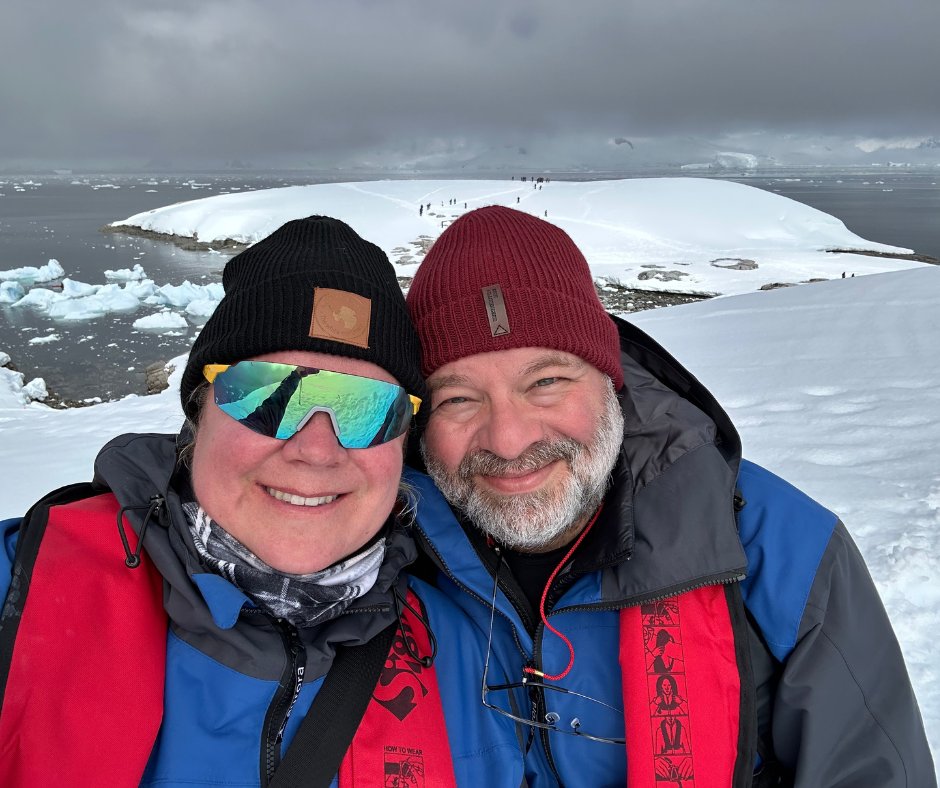 A smiling couple in expedition parkas on deck in Antarctica, icebergs and snowy peaks behind them.