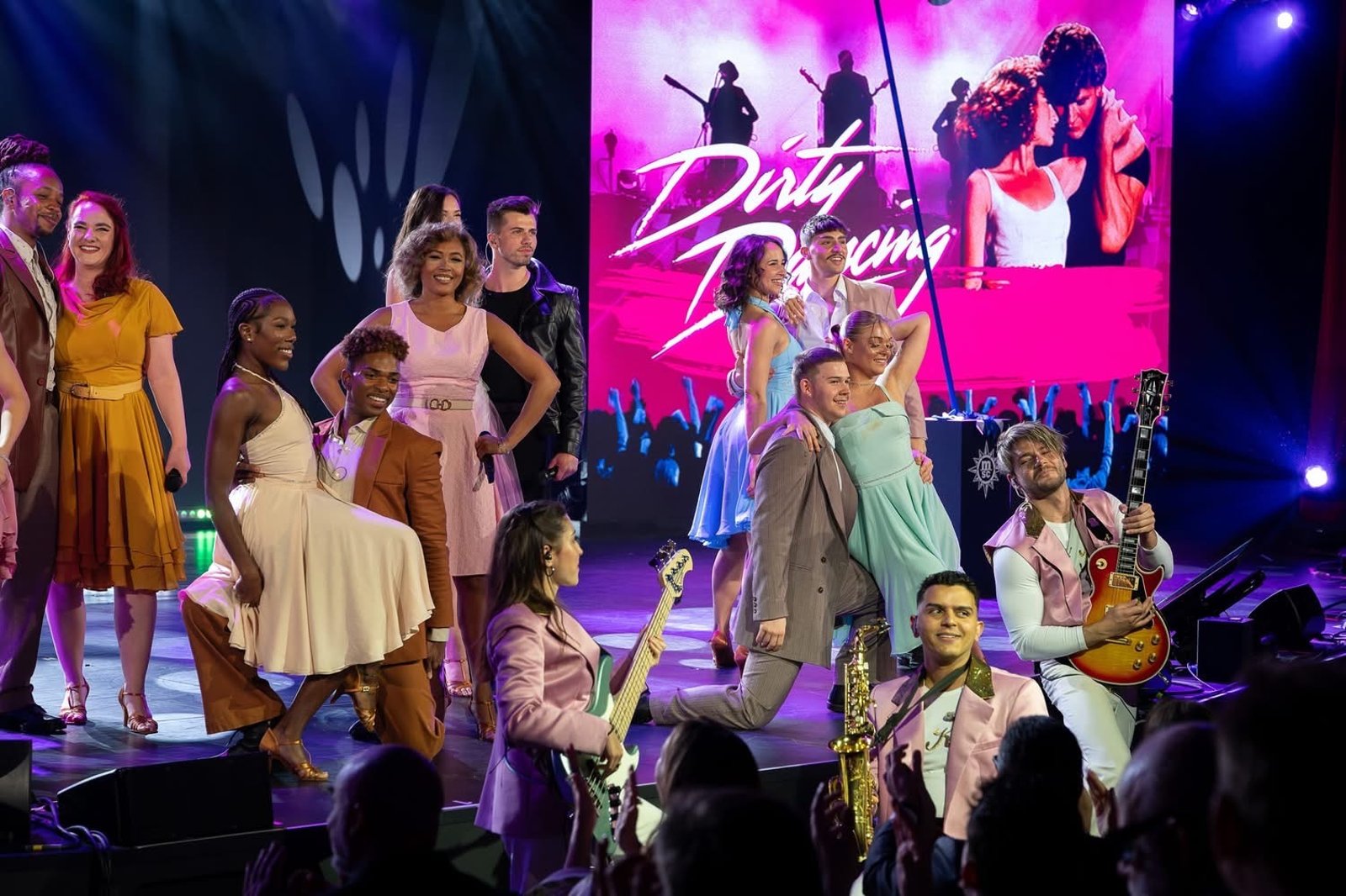 A cruise ship production cast in pastel costumes posing on a lit stage in front of a large Dirty Dancing screen.