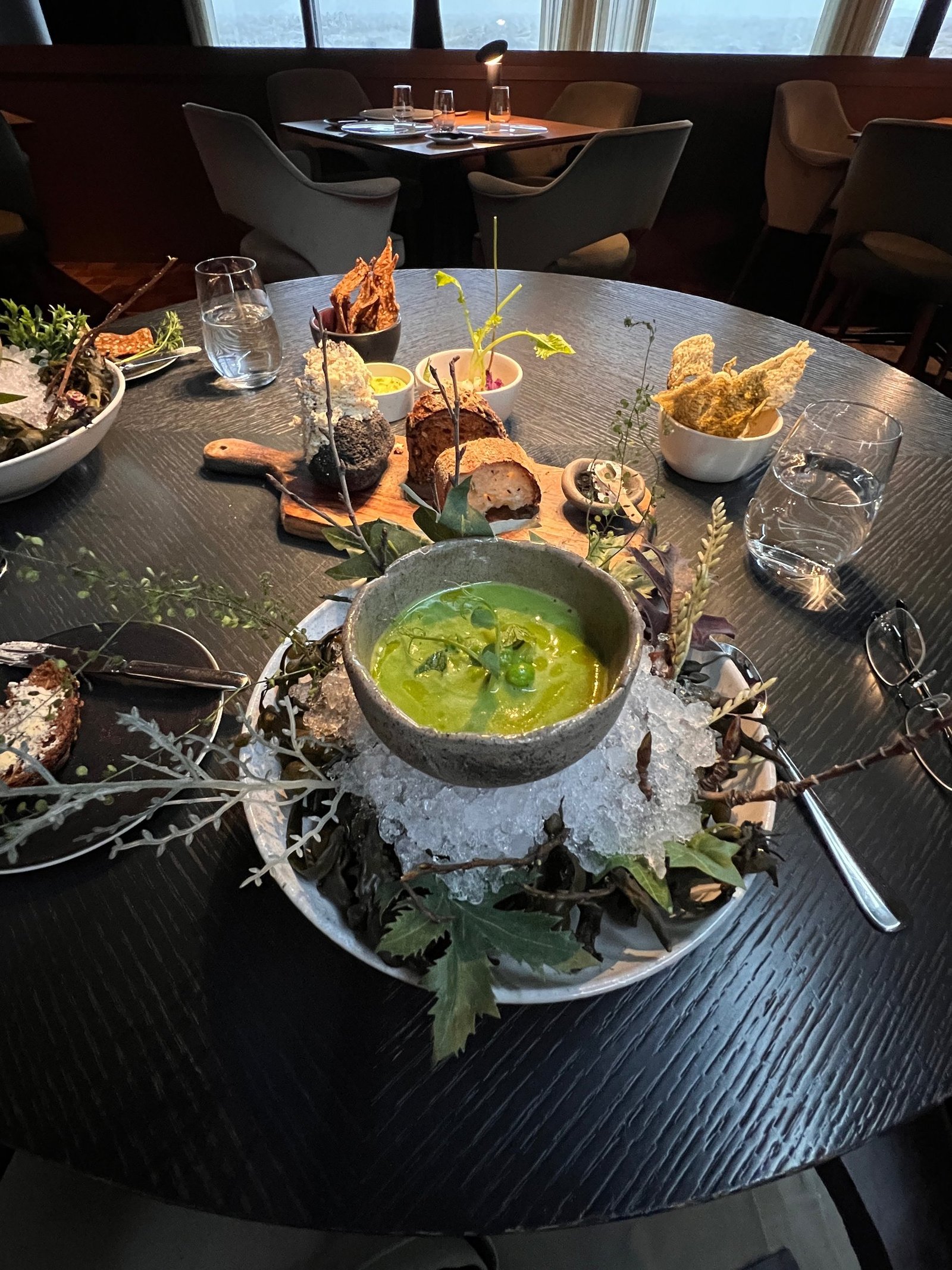A tasting-menu course of chilled green soup set on crushed ice with foraged herbs, breads and crisps on a dark table beside a panoramic window.