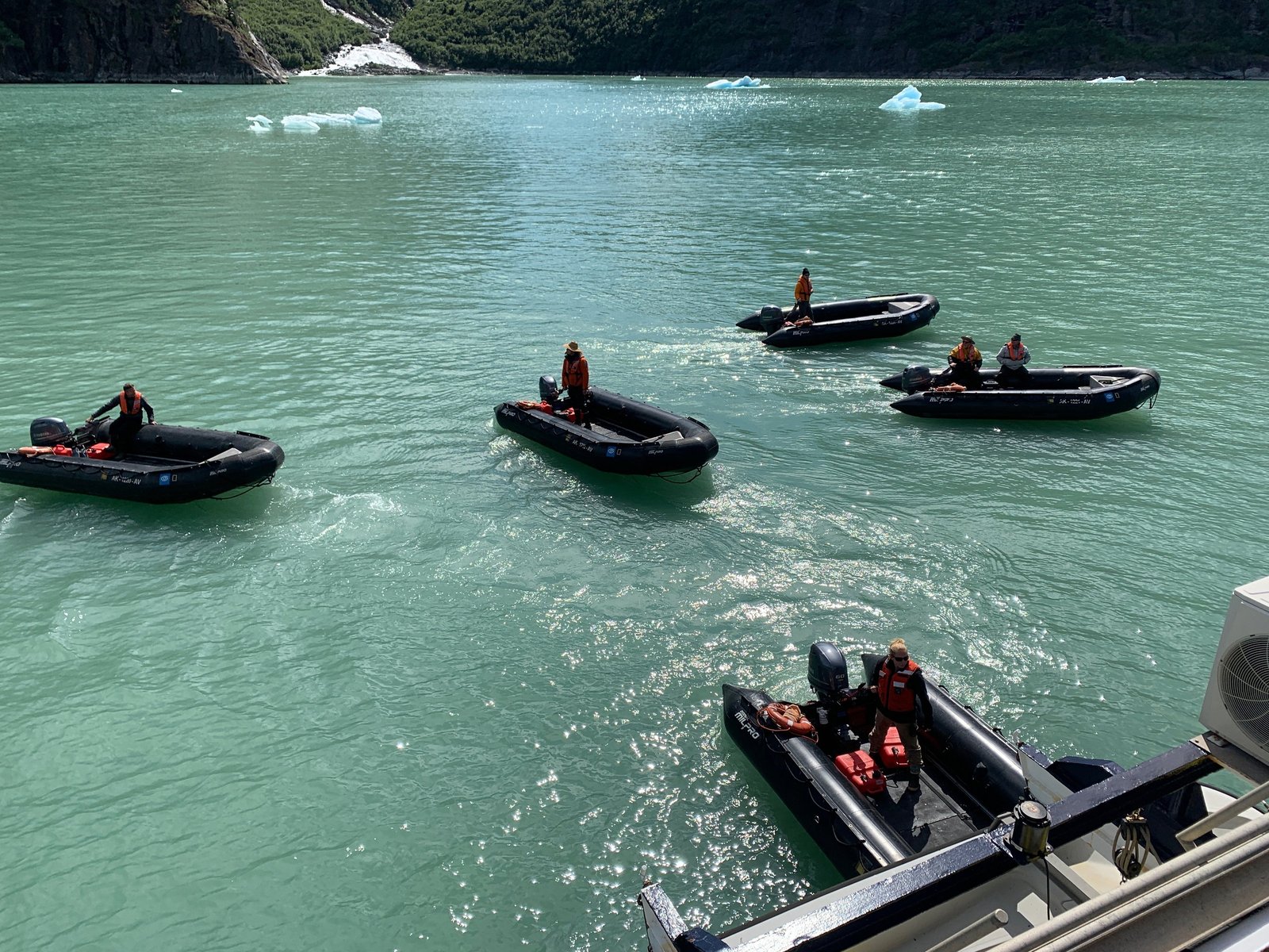 Five Zodiacs with drivers aboard idling on green glacial water beside an expedition ship, small icebergs drifting nearby.