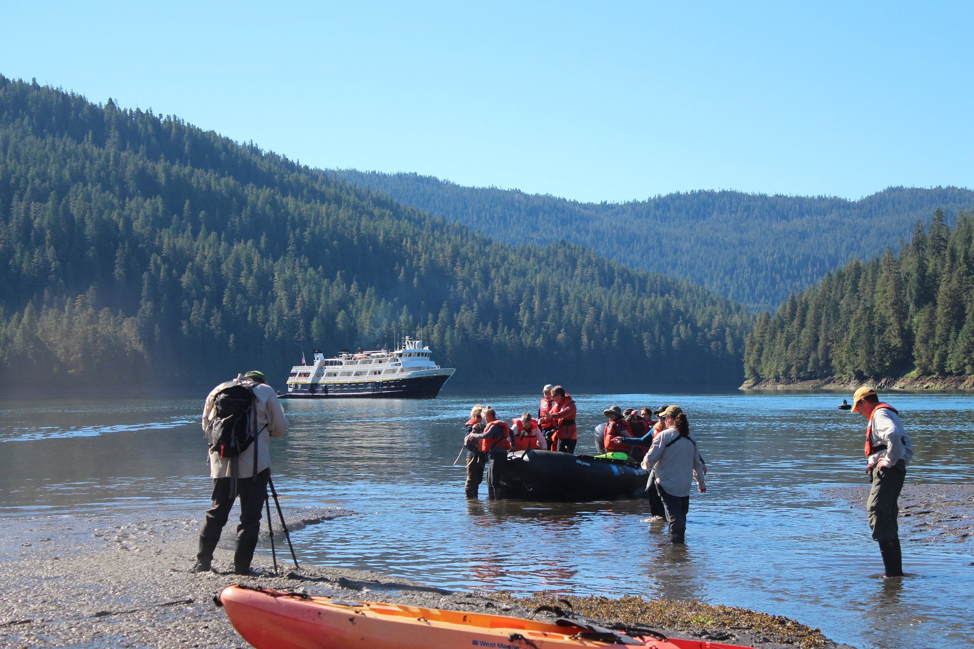 Tourists board a zodiac boat for an Alaskan cruise excursion near a large ship in a forested fjord.
