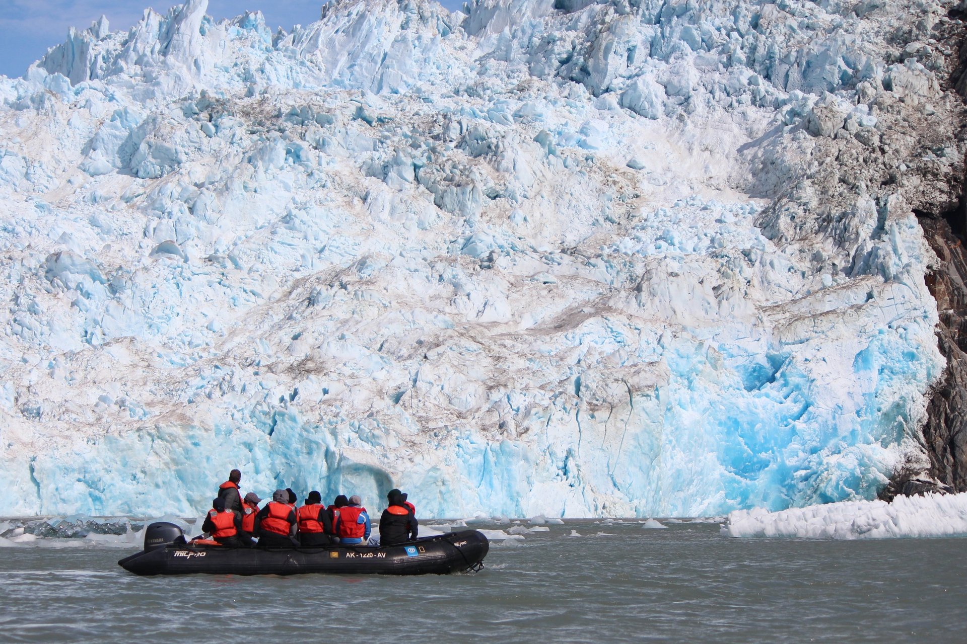 Tourists in a boat viewing the massive blue ice wall of a glacier in Alaska.