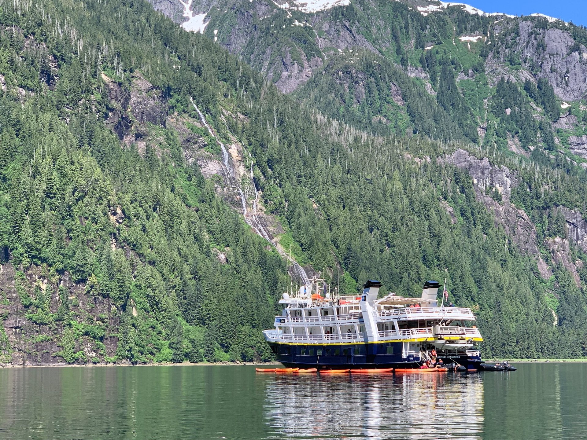 An Alaska expedition cruise ship anchored in a calm fjord near a waterfall and lush green mountains.