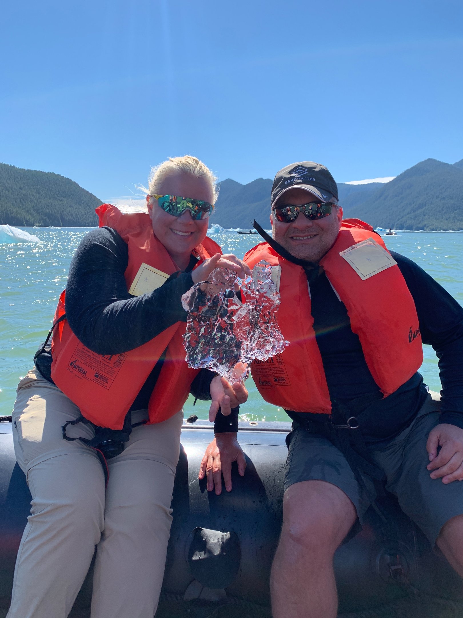 Couple on an Alaskan boat tour holding up a piece of glacial ice with mountains in the background.